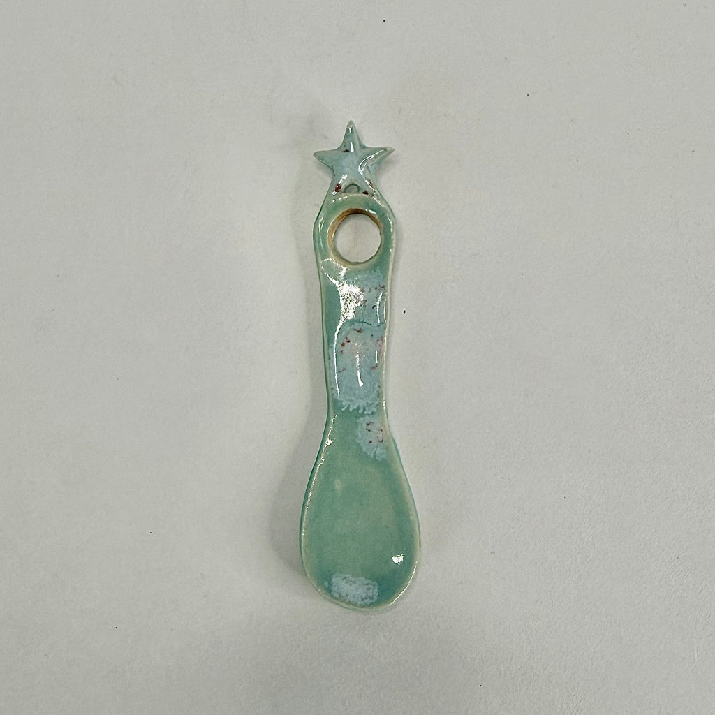 A small ceramic spoon-shaped holder with a star on top and a circular hole, glazed in a light mint green with white speckled accents, on a plain light gray background.