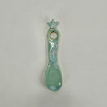 A small ceramic spoon-shaped holder with a star on top and a circular hole, glazed in a light mint green with white speckled accents, on a plain light gray background.