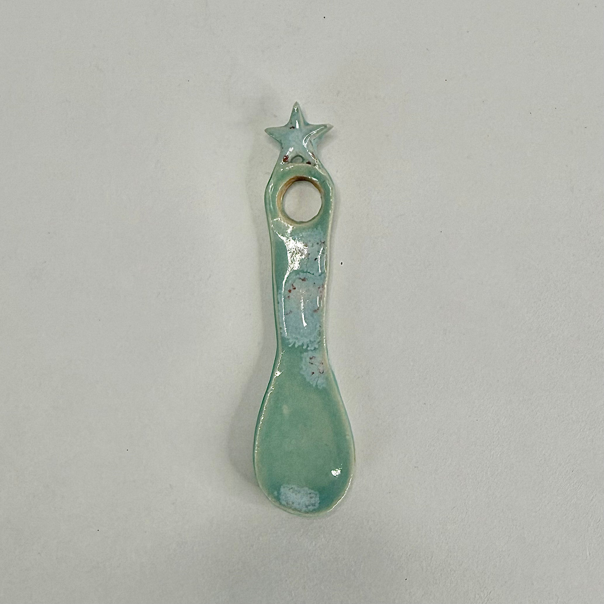 A small ceramic spoon-shaped holder with a star on top and a circular hole, glazed in a light mint green with white speckled accents, on a plain light gray background.