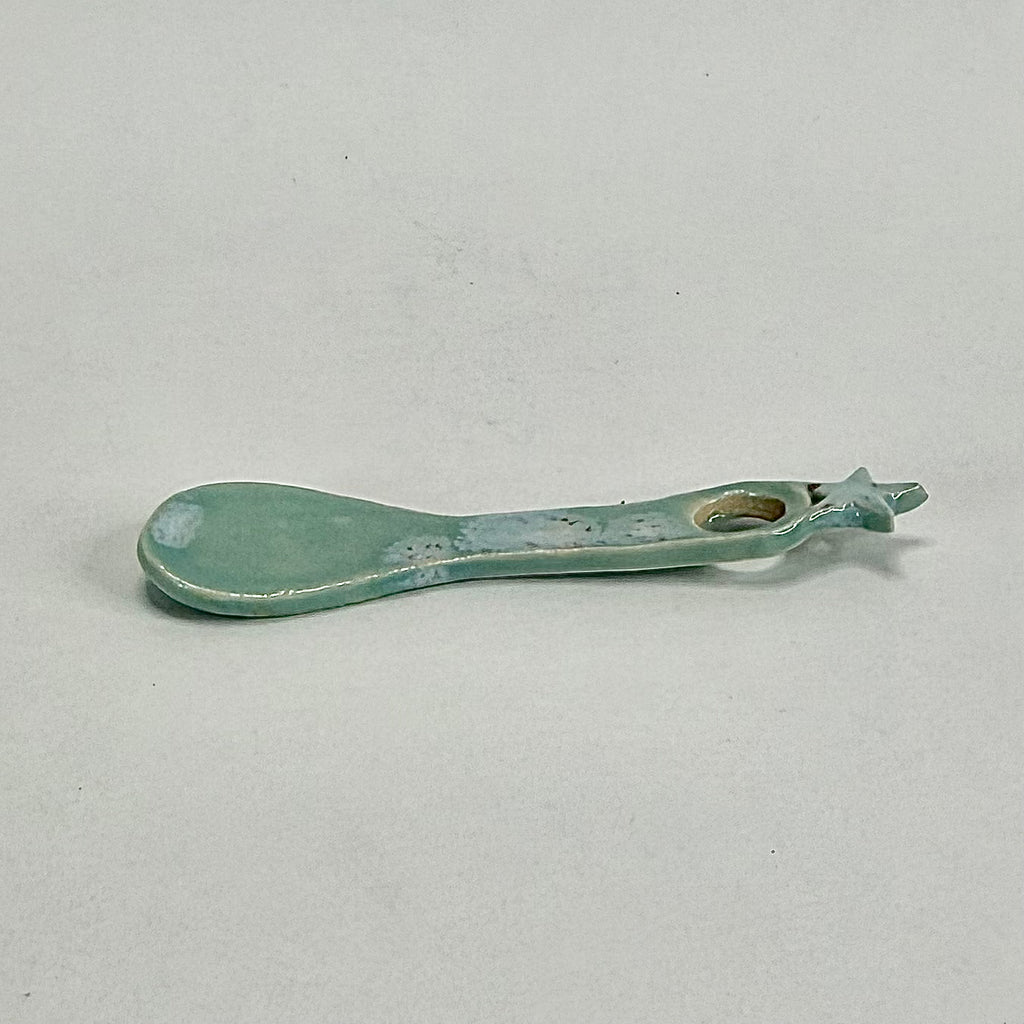 A small ceramic spoon or scoop with a rounded bowl and a looped handle, glazed in a pale mint green or aqua color with some distressed, speckled detailing, shown on a neutral light gray background.