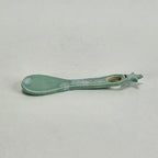 A small ceramic spoon or scoop with a rounded bowl and a looped handle, glazed in a pale mint green or aqua color with some distressed, speckled detailing, shown on a neutral light gray background.