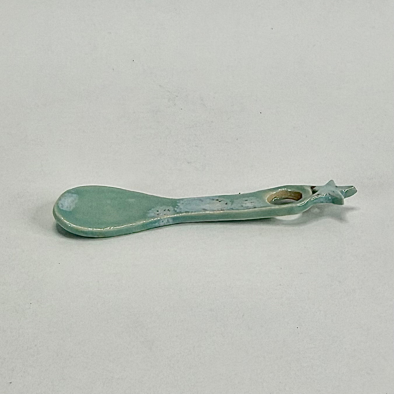 A small ceramic spoon or scoop with a rounded bowl and a looped handle, glazed in a pale mint green or aqua color with some distressed, speckled detailing, shown on a neutral light gray background.
