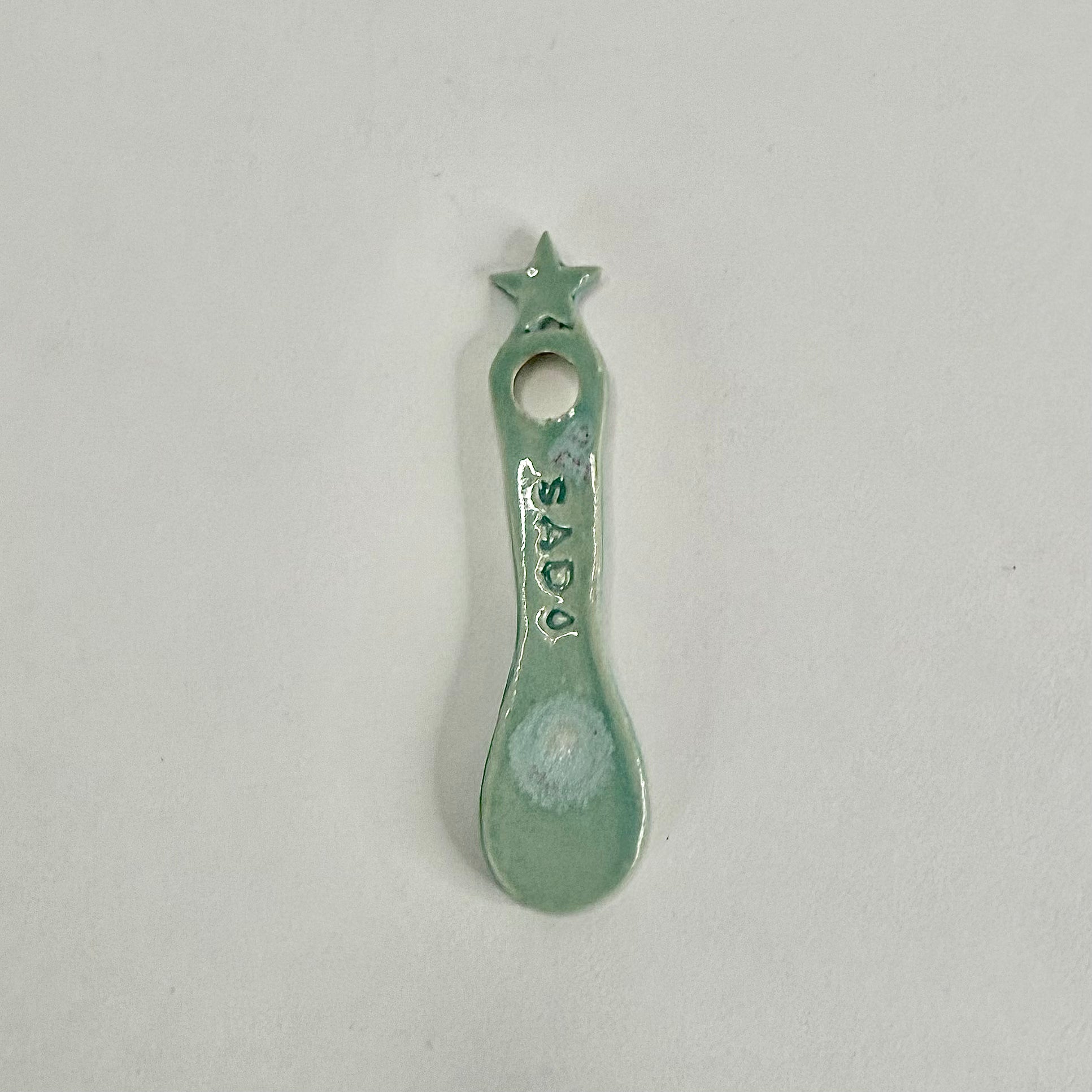 A small ceramic spoon or scoop with a star-shaped handle and a hole at the top, decorated with the name 'SAD' and a white circular mark near the bowl, shown on a plain light background.