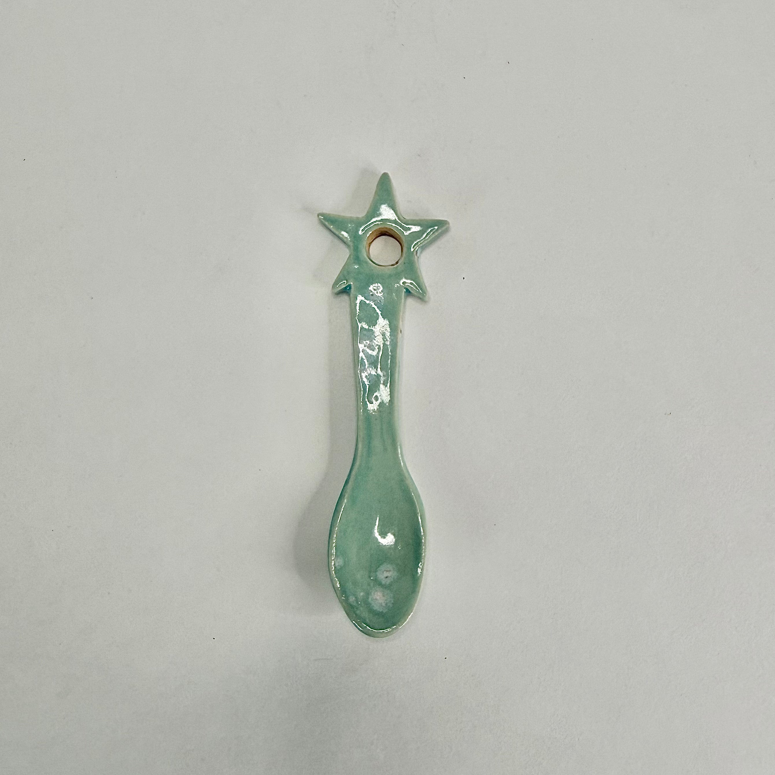 A small ceramic spoon-shaped ornament with a star handle and a hole at the top, glazed in a light mint green color, shown on a plain light gray background.