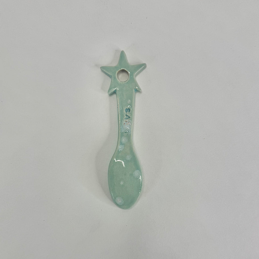 A small ceramic spoon-shaped ornament with a star handle, glazed in a pale mint green color with white speckled details, shown on a plain light gray background.