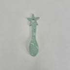 A small ceramic spoon-shaped ornament with a star handle, glazed in a pale mint green color with white speckled details, shown on a plain light gray background.