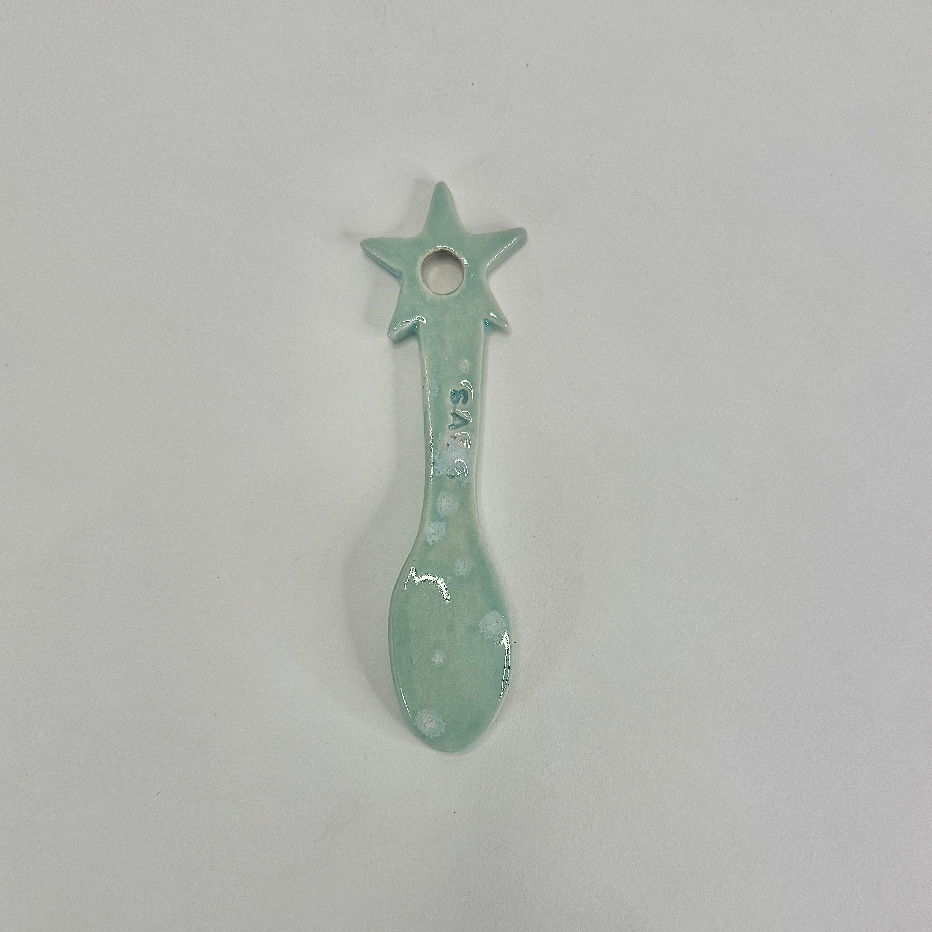 A small ceramic spoon-shaped ornament with a star handle, glazed in a pale mint green color with white speckled details, shown on a plain light gray background.