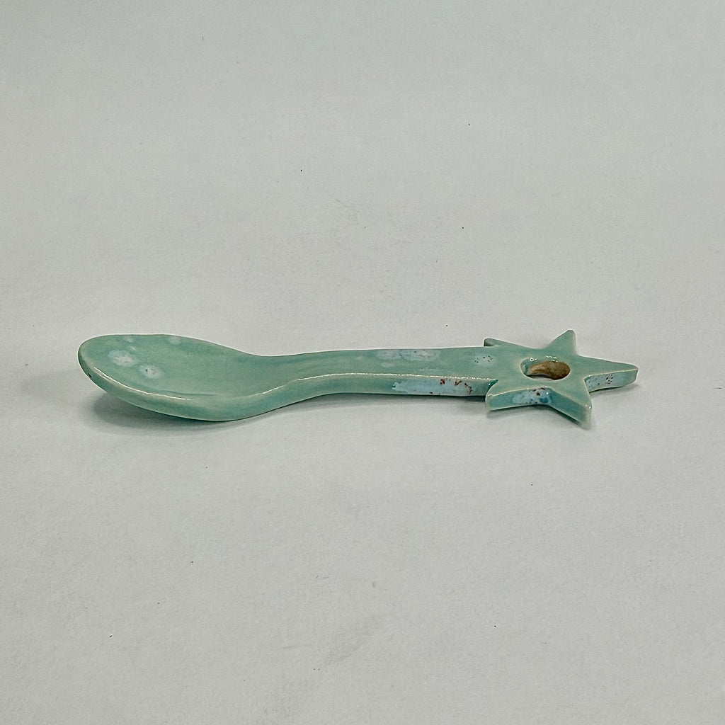 A small ceramic spoon-shaped object with a star handle, finished in a glossy light aqua glaze with some distressed blue and brown patina, shown on a neutral light gray background.