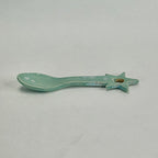 A small ceramic spoon-shaped object with a star handle, finished in a glossy light aqua glaze with some distressed blue and brown patina, shown on a neutral light gray background.
