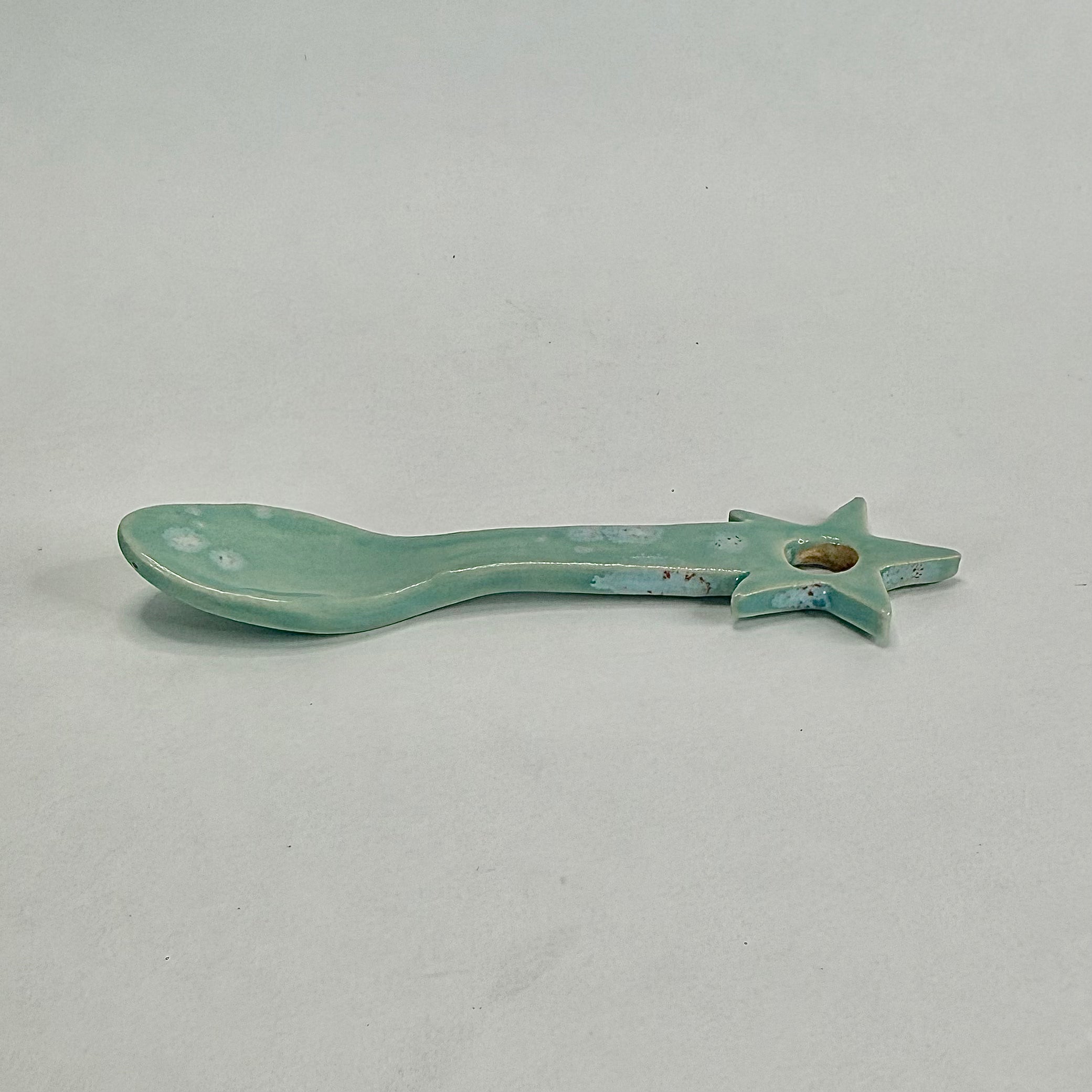 A small ceramic spoon-shaped object with a star handle, finished in a glossy light aqua glaze with some distressed blue and brown patina, shown on a neutral light gray background.