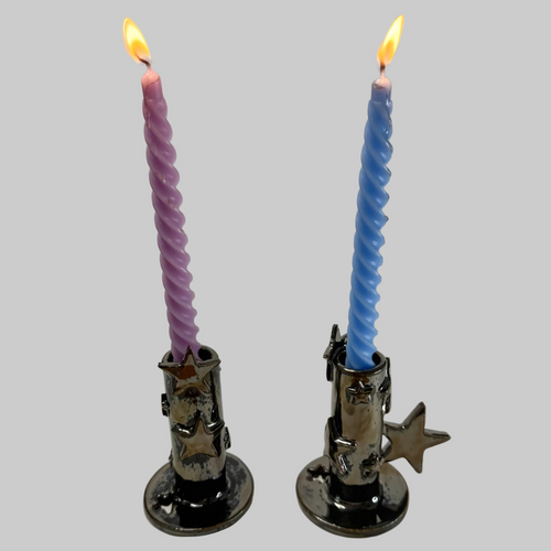 Two twisted taper candles, one purple and one blue, lit and displayed in silver candle holders with star details, set against a plain light gray background.