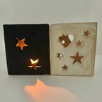 Two square ceramic candle holders with star and heart cutouts, one black and one cream-colored, shown lit with warm orange light on a white surface.