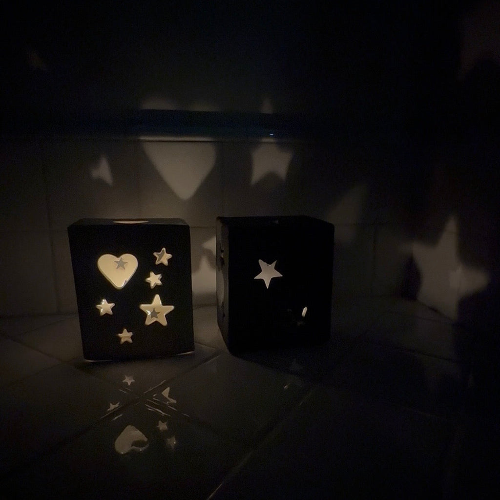 Two black cube-shaped decorative lanterns with star and heart cutouts, casting star-shaped shadows on a dark tiled wall and floor.