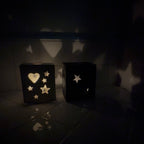 Two black cube-shaped decorative lanterns with star and heart cutouts, casting star-shaped shadows on a dark tiled wall and floor.
