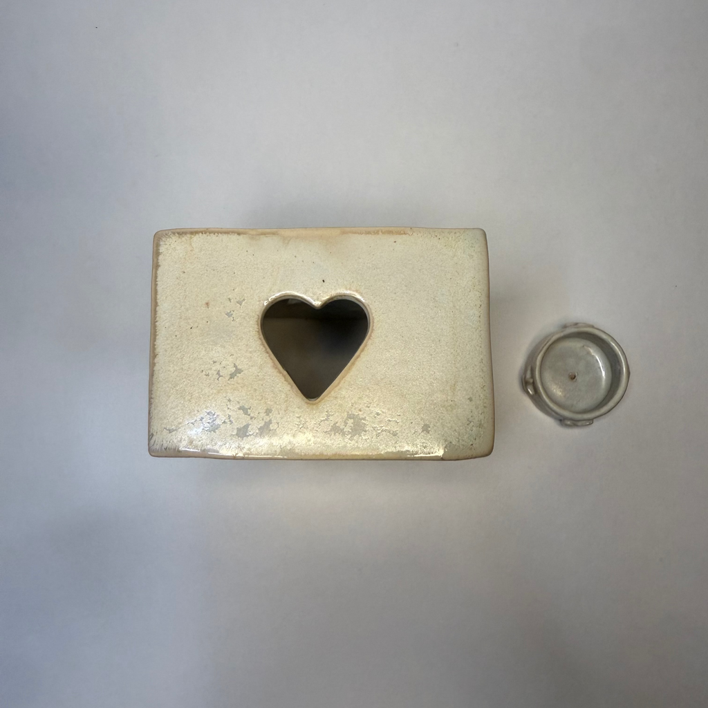 A square cream-colored ceramic container with a heart-shaped cutout on the front, shown next to its small round base on a neutral gray background.