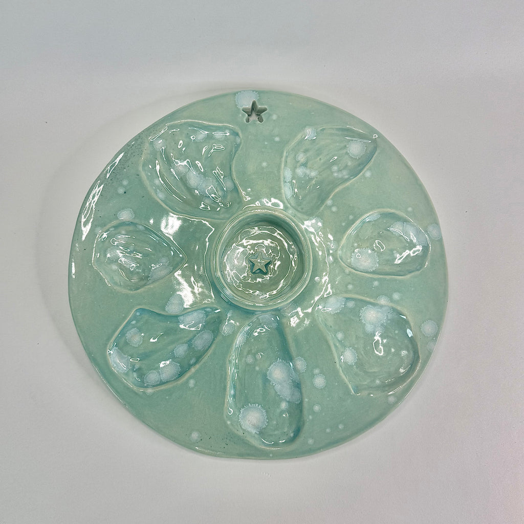 A round, glossy ceramic plate with oyster shape cut outs with a light mint green glaze and raised mini bowl for mignottes and sauces, photographed from above on a plain light background