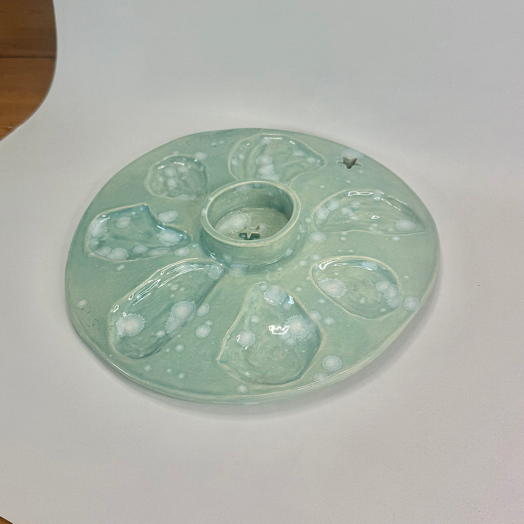 A round ceramic artist palette with multiple shallow wells and a central well, glazed in a pale mint green with white bubbly speckling, photographed on a white studio surface.