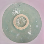 The back of a round ceramic plate with a glossy pale mint green glaze, featuring embossed star details around a raised cream-colored center ring and small white speckles scattered across the surface, shown from a top-down view on a white background.