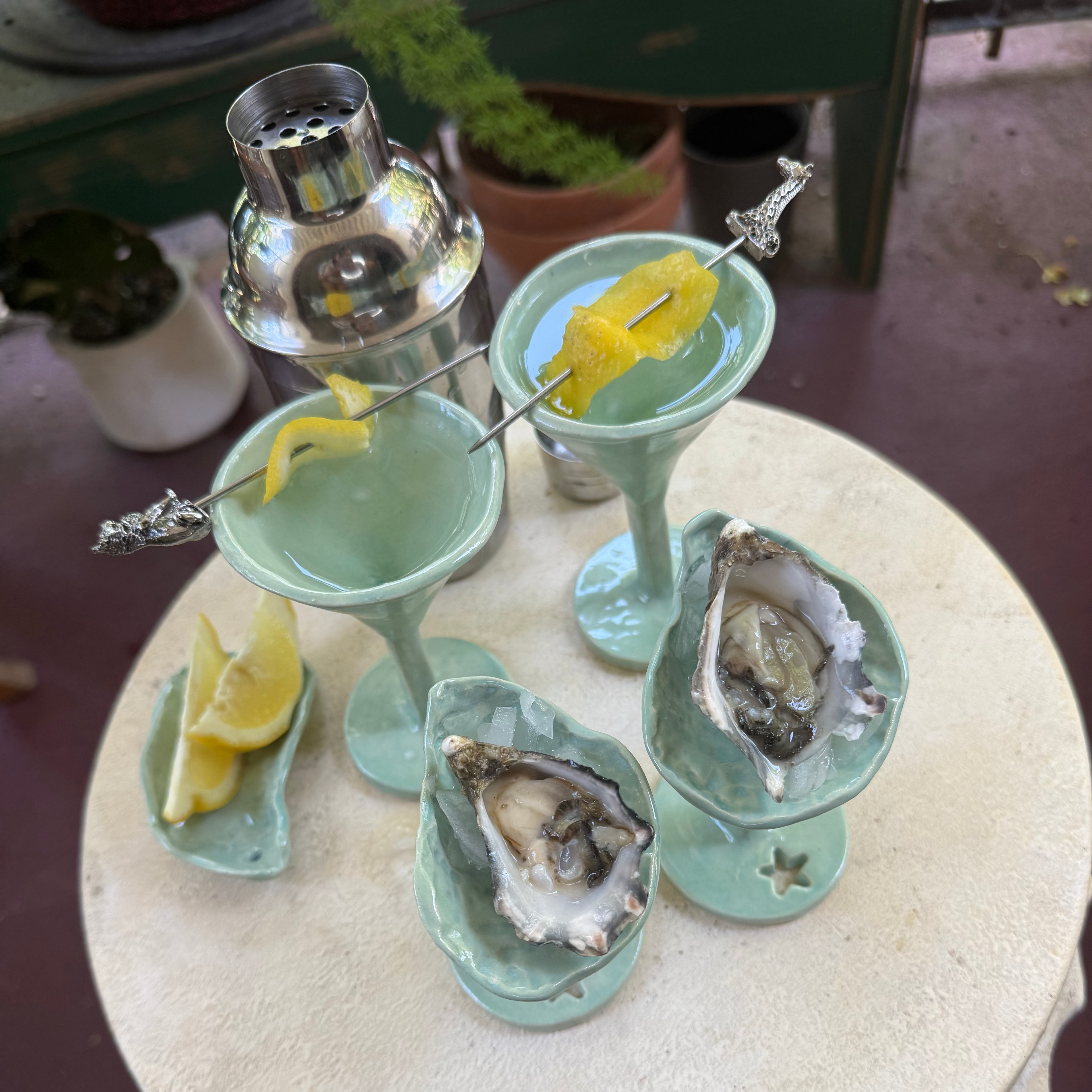 A styled food and drink presentation on a round table, featuring two pale aqua martini glasses with lemon rinds, two oysters in shell dishes, a cocktail shaker, and lemon wedges on a patio-style surface.