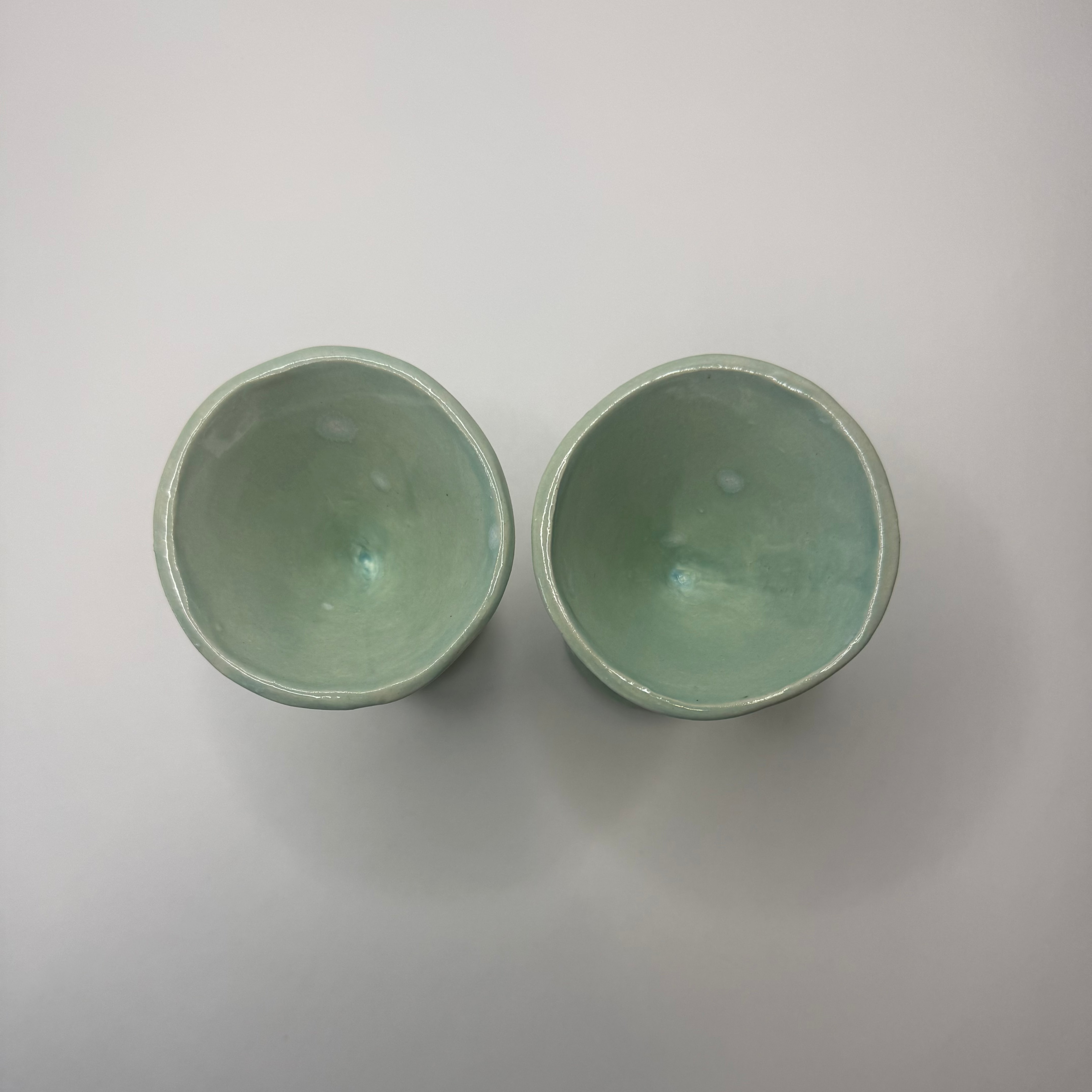 Two small handmade ceramic cups with a glossy pale green glaze, shown from a top-down view on a neutral light gray background.