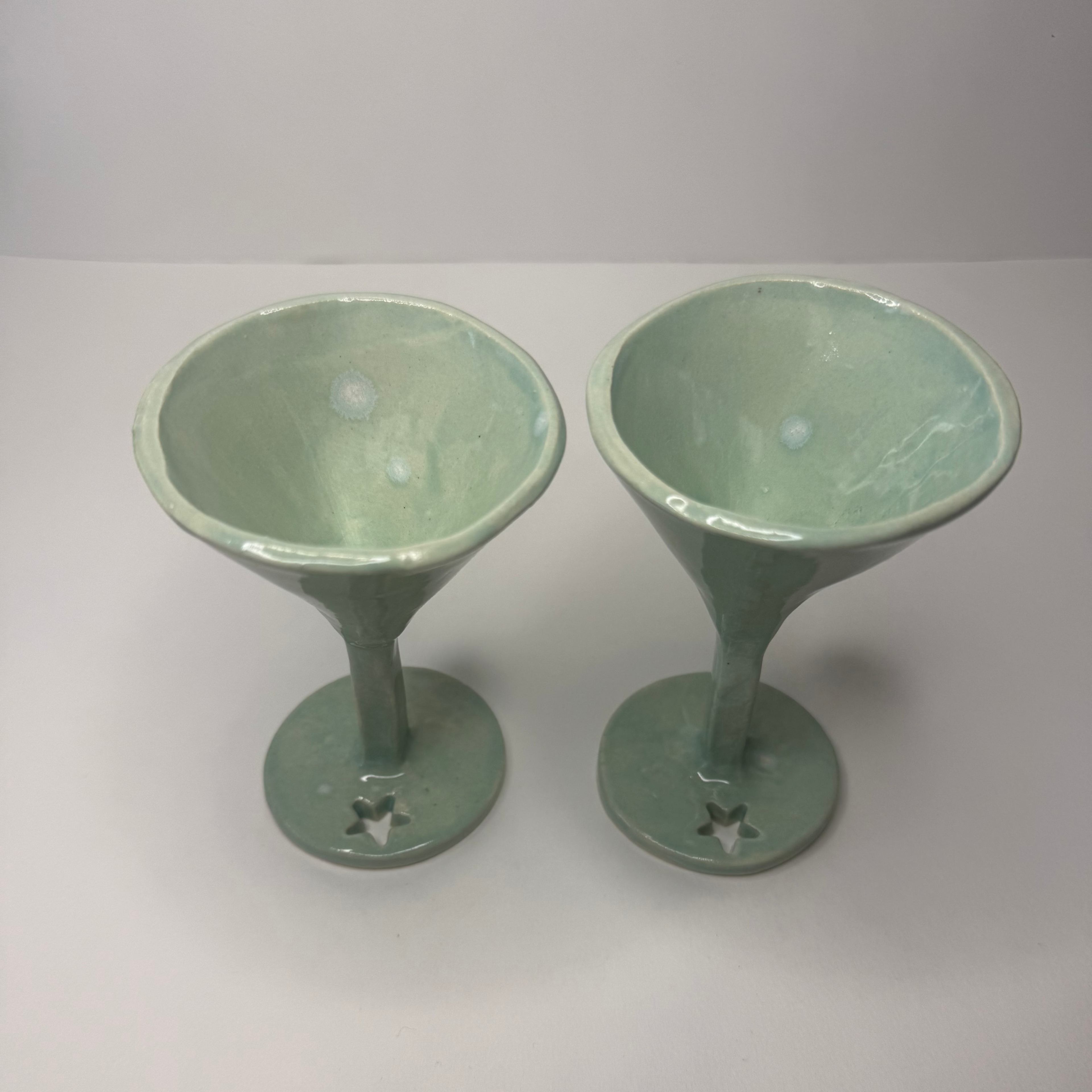 A pair of handmade ceramic stemmed goblets with a glossy light green glaze, featuring star-shaped cutouts at the base of the circular feet, set against a plain light gray background.