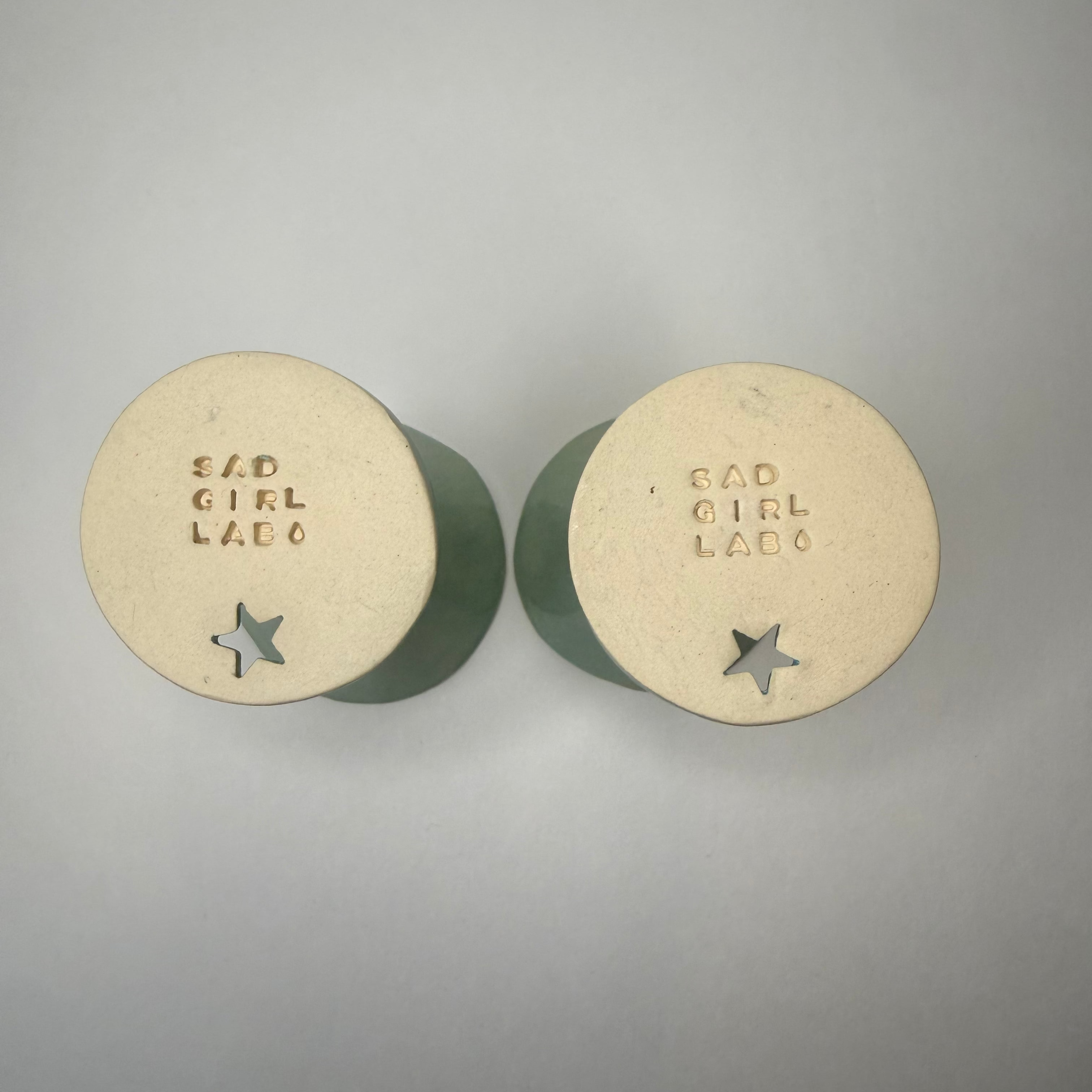 Two round ceramic bases with the words 'SAD GIRL LAB' and a star cutout, shown from the bottom on a neutral gray background.